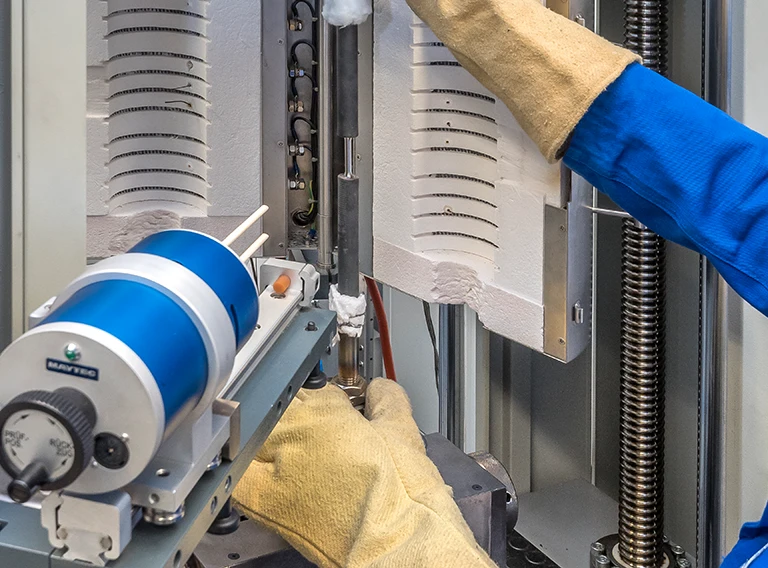 Eine Person, die Schutzhandschuhe und einen blauen Laborkittel trägt, bedient ein wissenschaftliches Gerät, möglicherweise eine Materialprüfmaschine, bei der Metall- und isolierte Komponenten sichtbar sind.