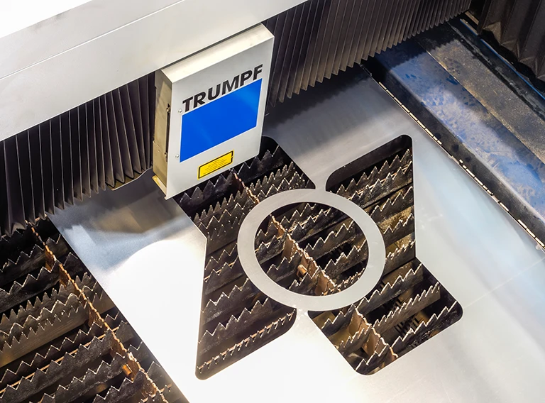 Eine TRUMPF Laserschneidmaschine schneidet präzise eine kreisförmige Form aus einem Blech, unter der die gezackten Stützen sichtbar sind.