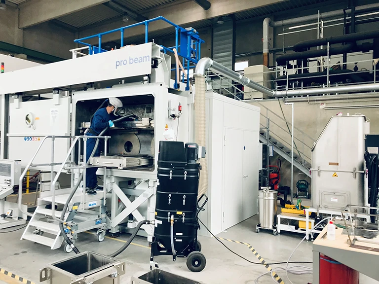 Eine Person in Schutzkleidung bedient eine große Industriemaschine mit der Aufschrift Pro Beam in einer sauberen, modernen Fabrik, in der verschiedene Geräte, Kabel und Lagereinheiten sichtbar sind.