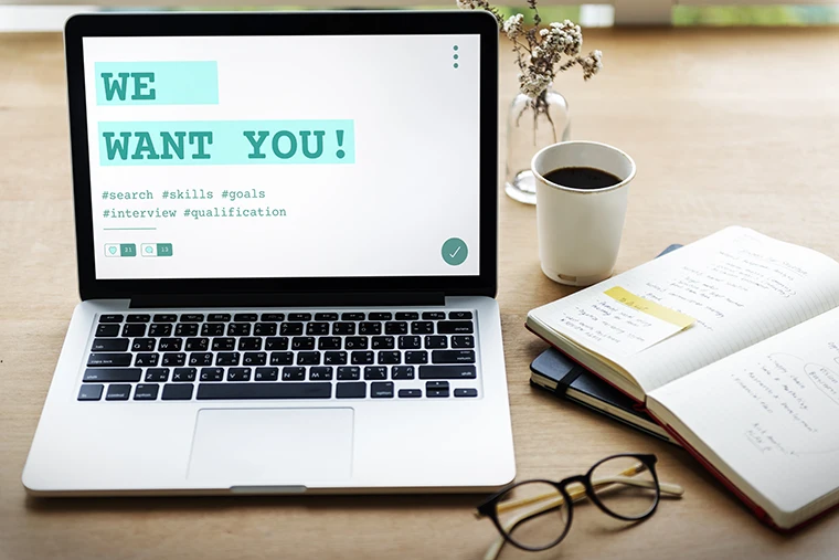 Auf einem hölzernen Schreibtisch steht ein Laptop mit der Aufschrift WE WANT YOU! und Hashtags. In der Nähe stehen eine Tasse Kaffee, ein Notizbuch mit handschriftlichen Notizen und eine Brille.