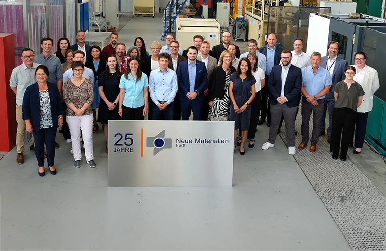 Eine Gruppe von etwa 30 Personen steht in einer Fabrik- oder Industriehalle hinter einem Schild mit der Aufschrift 25 Jahre Neue Materialien Fürth, das das 25-jährige Bestehen des Unternehmens feiert.
