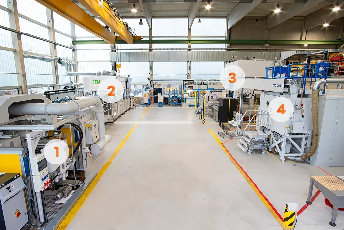 Eine helle, moderne Industrieanlage mit großen Maschinen, die mit 1 bis 4 gekennzeichnet sind, hohen Decken, Sicherheitsbarrieren, einem gelben Kran und großen Fenstern an den Wänden, die natürliches Licht hereinlassen. Der Boden ist sauber und deutlich gekennzeichnet.