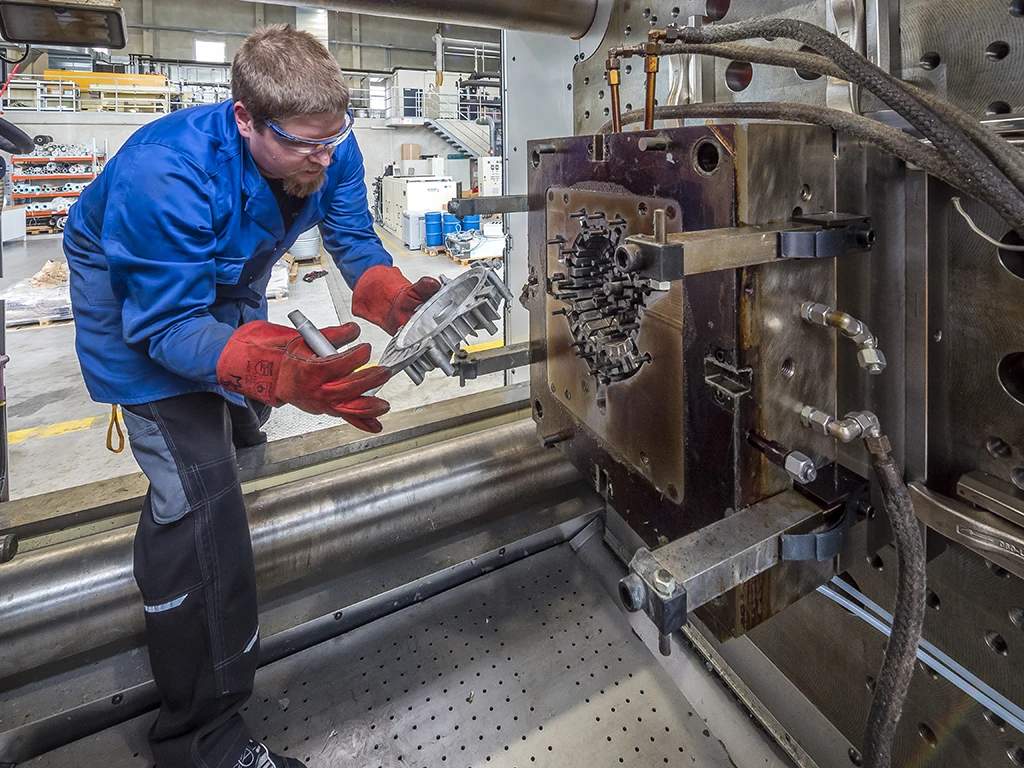 Ein Arbeiter mit Schutzhandschuhen und Schutzbrille entnimmt in einer Fabrik ein Metallteil aus einer Industriemaschine und prüft das geformte Bauteil genau.