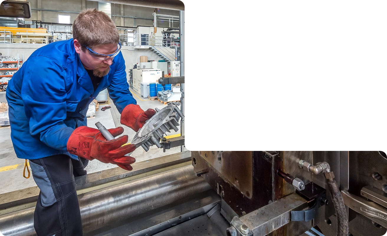Ein Mann mit Schutzbrille, blauem Mantel und roten Handschuhen hält ein Metallteil neben einer Industriemaschine in einer Fabrikumgebung. Ein Teil des Bildes ist geschwärzt, um einige Geräte zu verdecken.