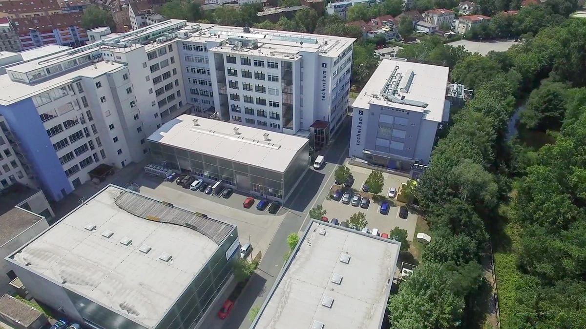 Luftaufnahme eines modernen Krankenhauskomplexes mit mehreren weißen Gebäuden, einem Parkplatz mit Autos und grünen Bäumen, die die Anlage an einem sonnigen Tag umgeben.